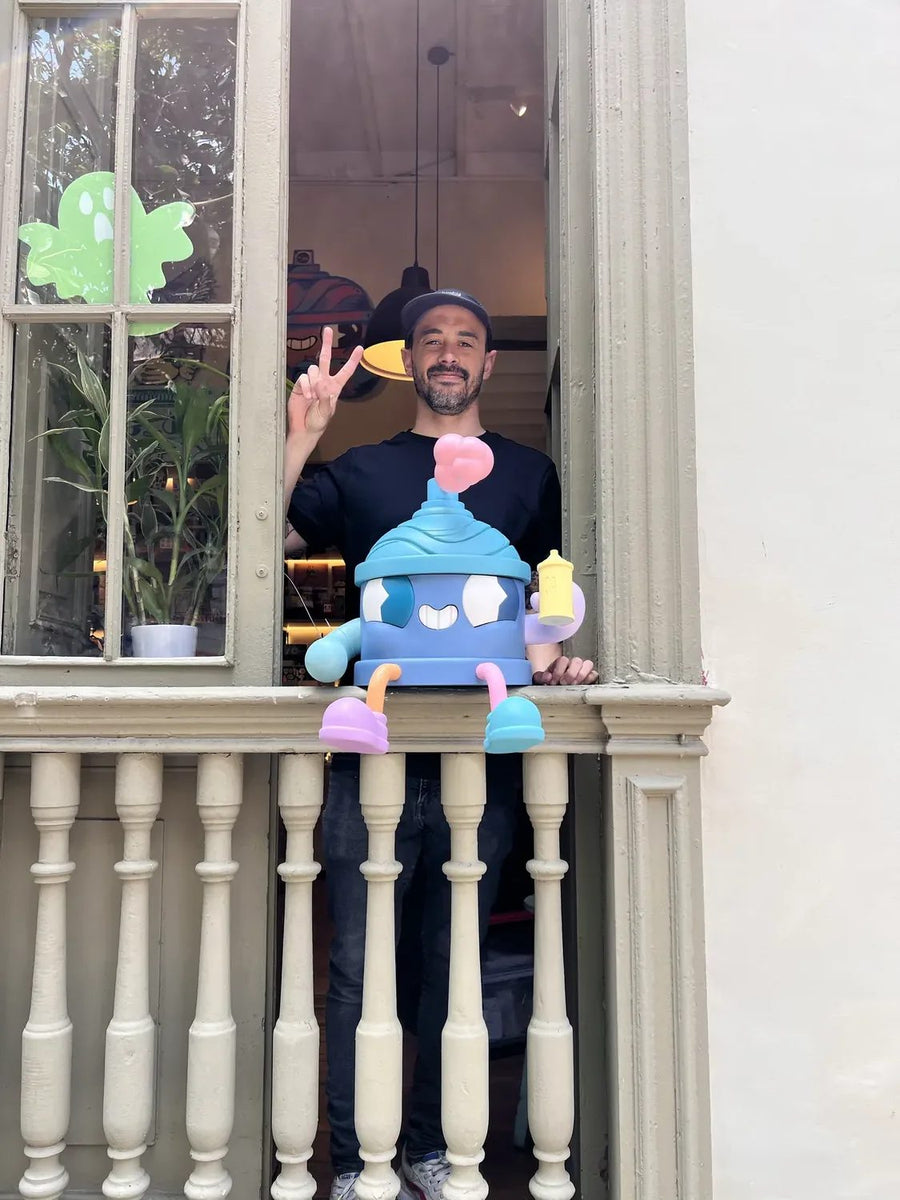 Uno de los artistas saluda desde la ventana del local con un art toy en la baranda
